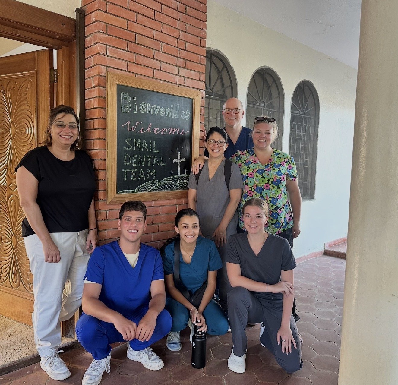 Smiles Bring Hope team gathered for a dental mission, posing beside a hand-lettered welcome chalkboard featuring a cross